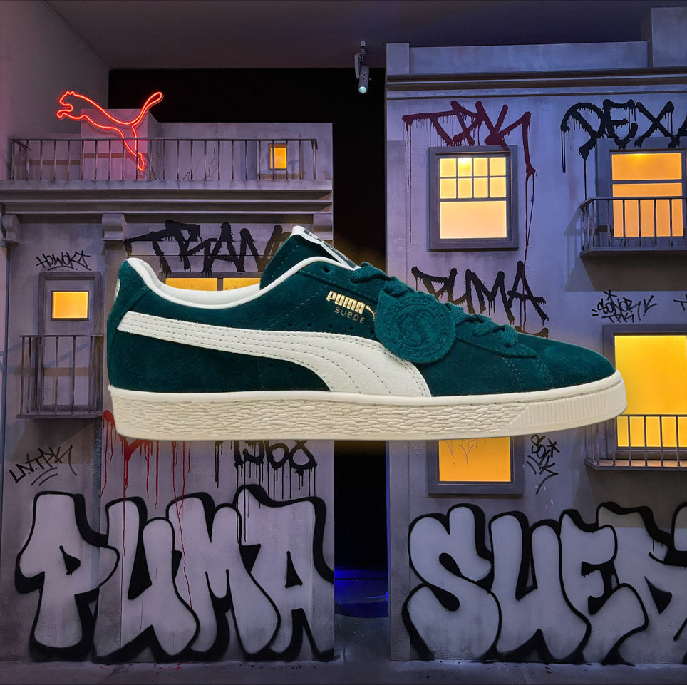A Puma Suede sneaker in dark teal green suede with a white side stripe and cream sole, shown floating in front of a graffiti-covered building backdrop with neon Puma branding.